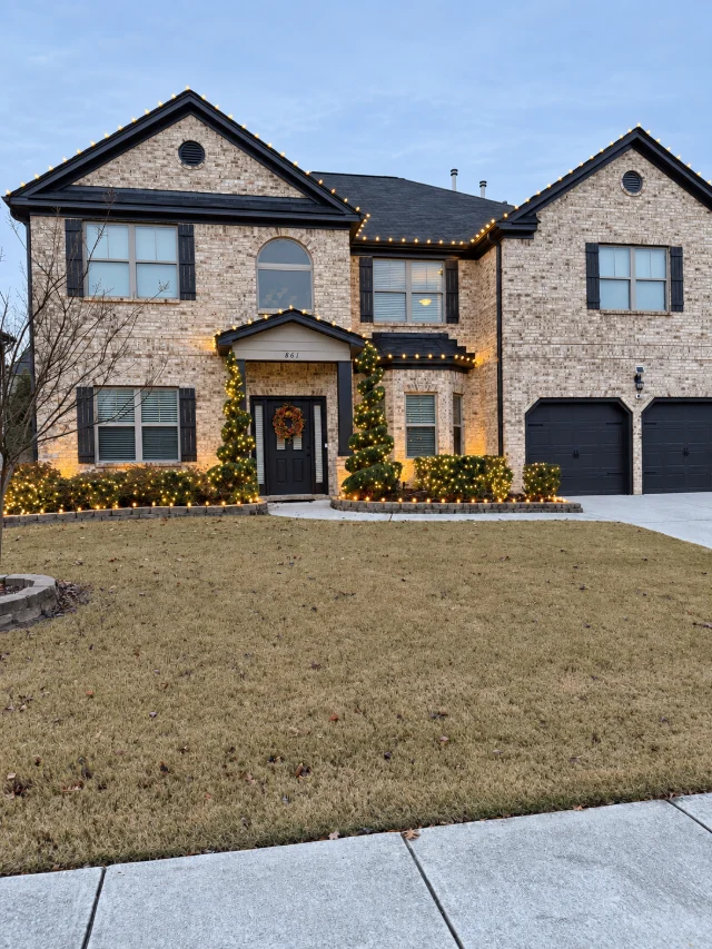 A daytime view of warm white roofline lights, wrapped evergreens, and shrub lighting.