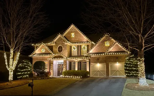 Brick-and-stone home at night with warm white roofline lights, illuminated wreaths, and lights around front-yard trees and shrubs.