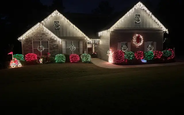 Home at night with white icicle-style lights along the rooflines, illuminated wreaths, and red and green lights on front shrubs.
