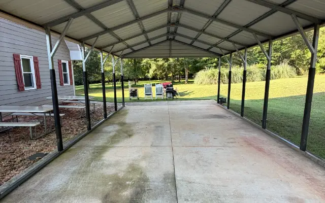 Covered concrete parking area beneath a metal carport beside a home.