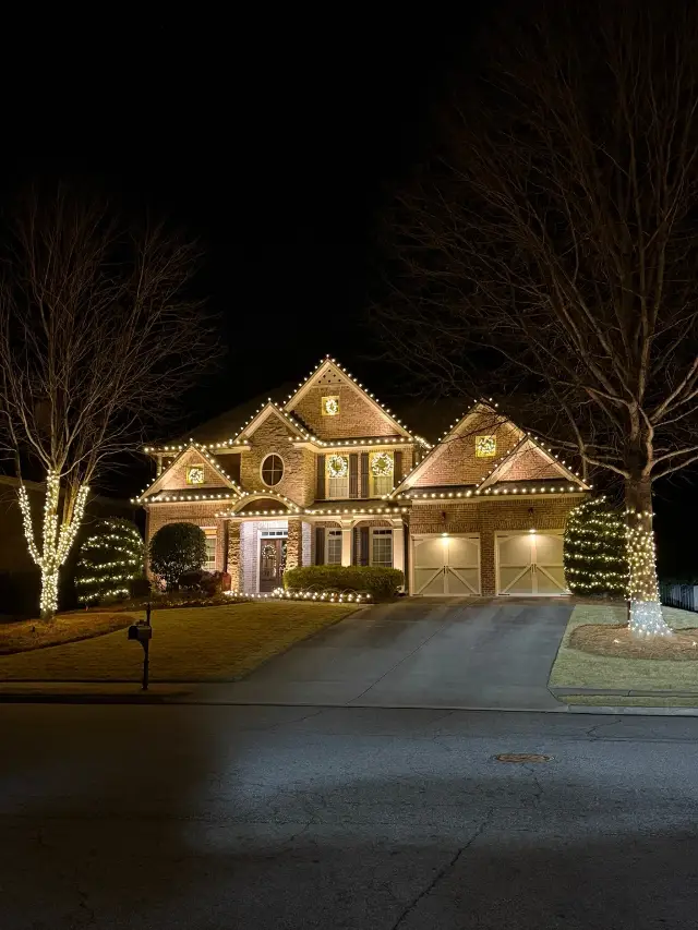 Brick-and-stone home at night with warm white roofline lights, illuminated wreaths, and lights around front-yard trees and shrubs.
