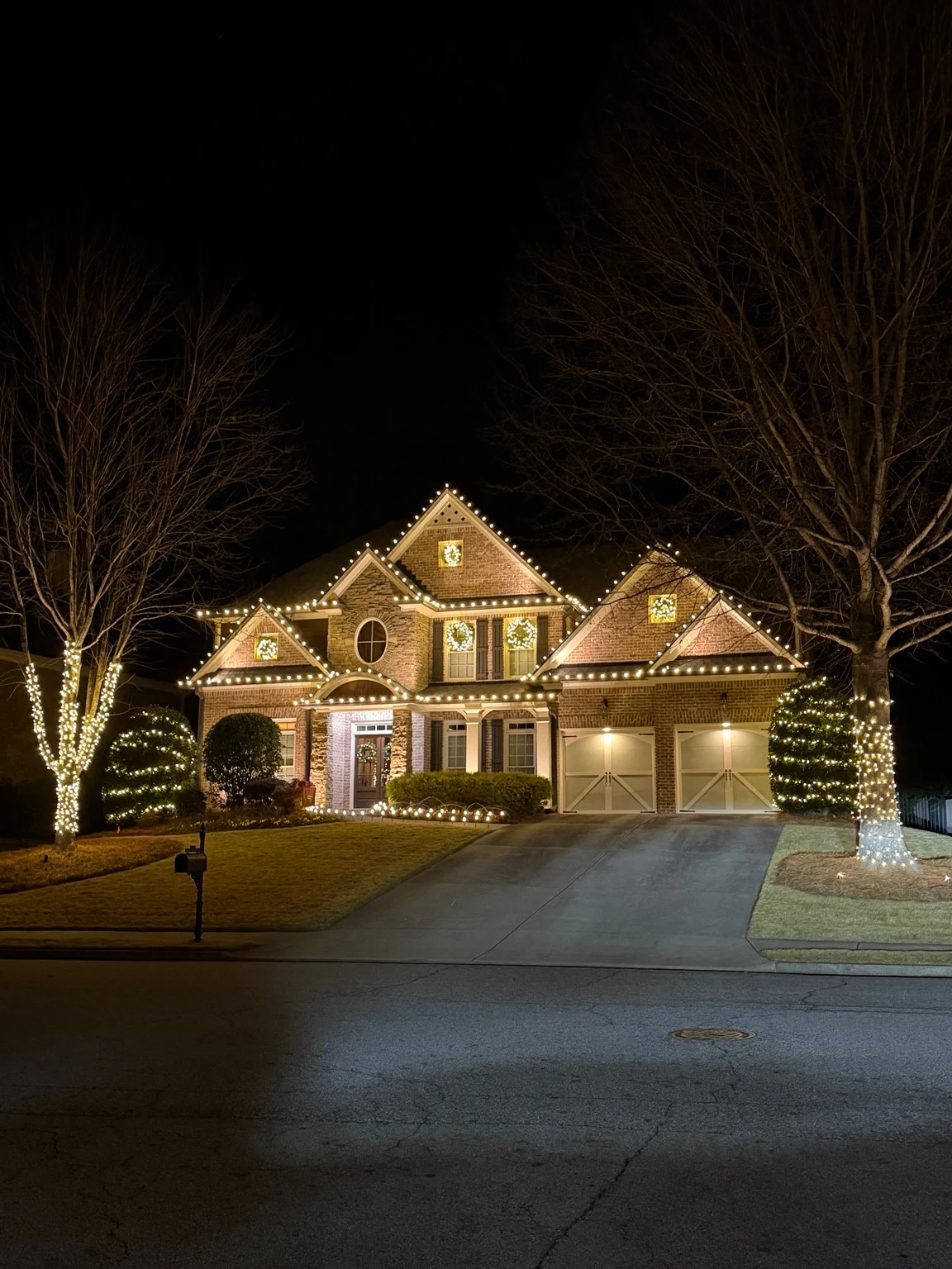 Brick-and-stone home at night with warm white roofline lights, illuminated wreaths, and lights around front-yard trees and shrubs.